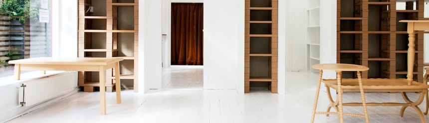 白い床に椅子とテーブルが並び壁際に木材で作られたおしゃれなデザインの棚が並んでいる横長の画像